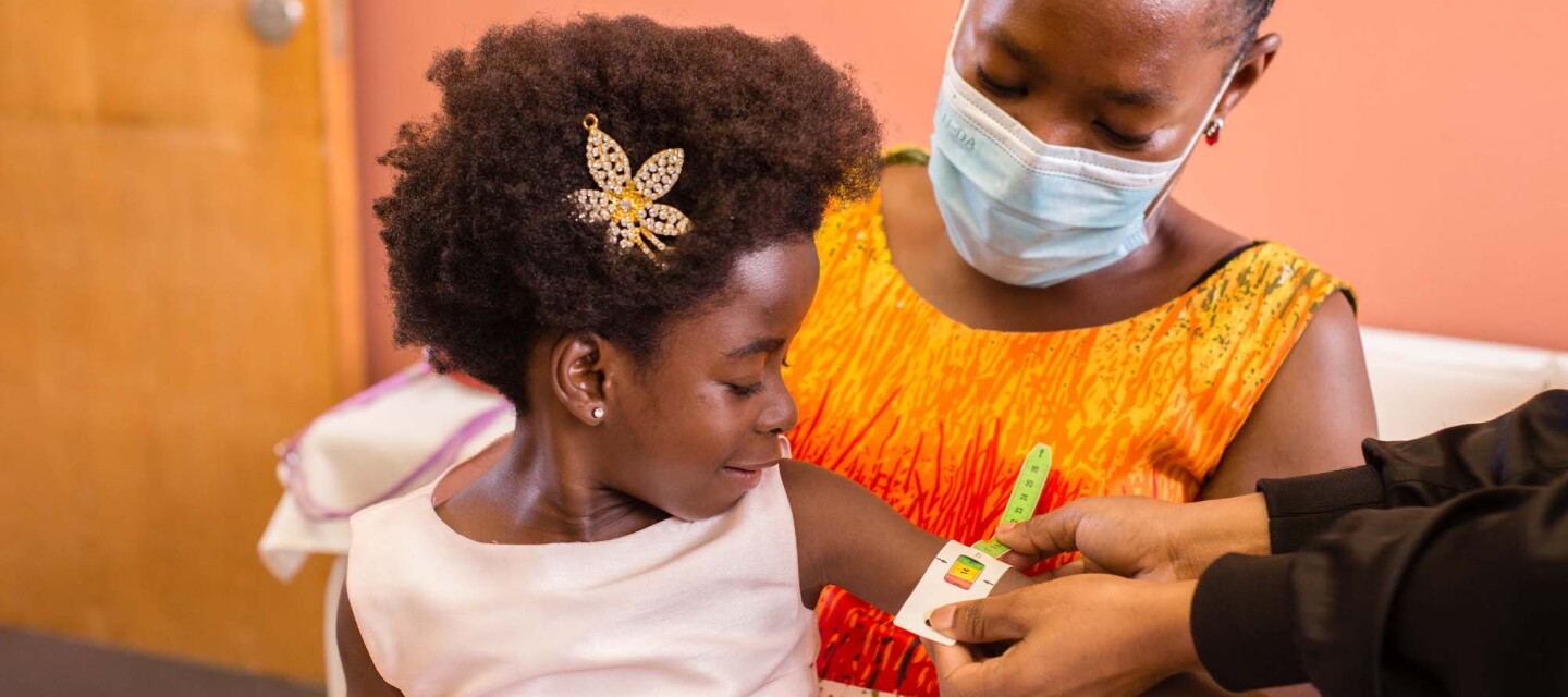 A girl sits on her mother's lap while having her arm measured by a healthcare professional.