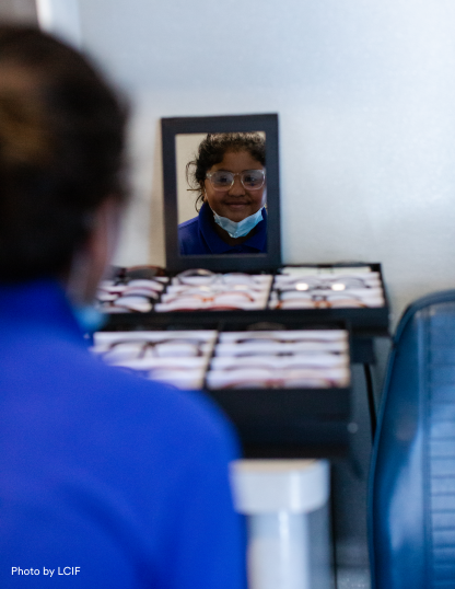 A girl looks into a mirror with a trial pair of eye glasses and smiles